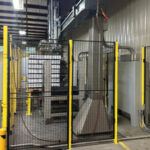 Side view of a cold storage palletizing system, featuring a gantry robot stacking product cases, surrounded by safety fences in an industrial environment.