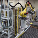 Automated machine tending system with a FANUC robot arm picking up carpet tiles from a conveyor line, preparing them for press loading in a manufacturing facility.